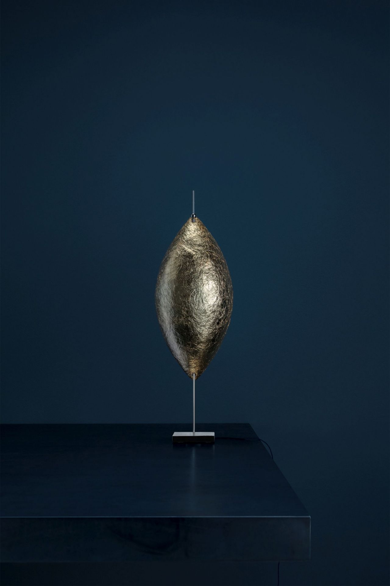 Malagolina Tischleuchte von Catellani & Smith in Gold: Moderne Lampe mit Blattgold-Effekt auf dunklem Tisch.