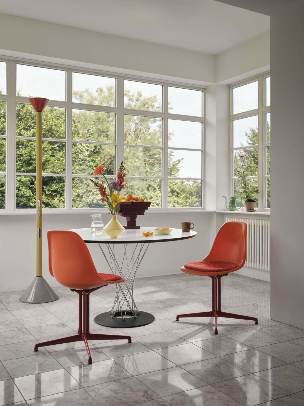 Esszimmer mit rundem Esstisch, Design-Stühlen in Orange und moderner Stehlampe vor großen Fenstern.