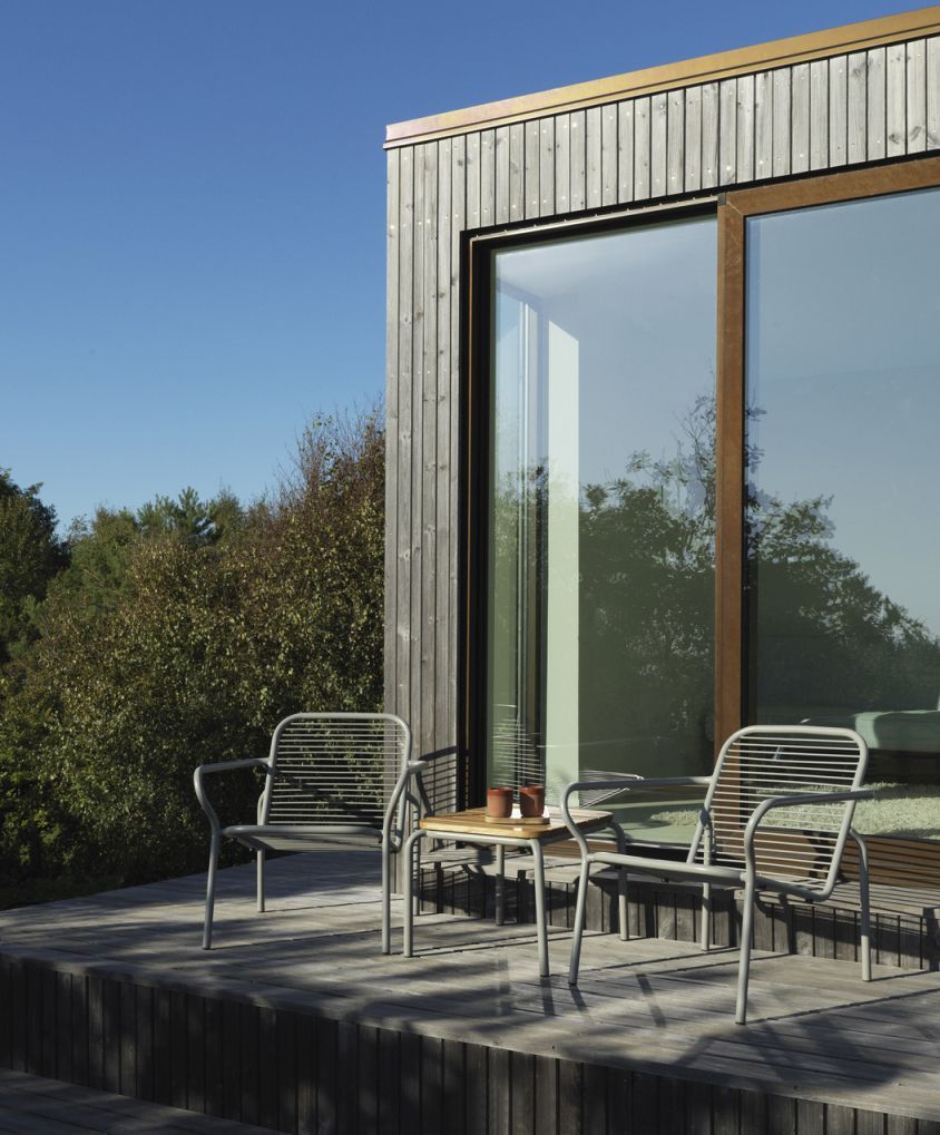 Normann Copenhagen Vig Table Outdoor mit zwei Stühlen auf einer Holzterrasse vor einem modernen Haus.