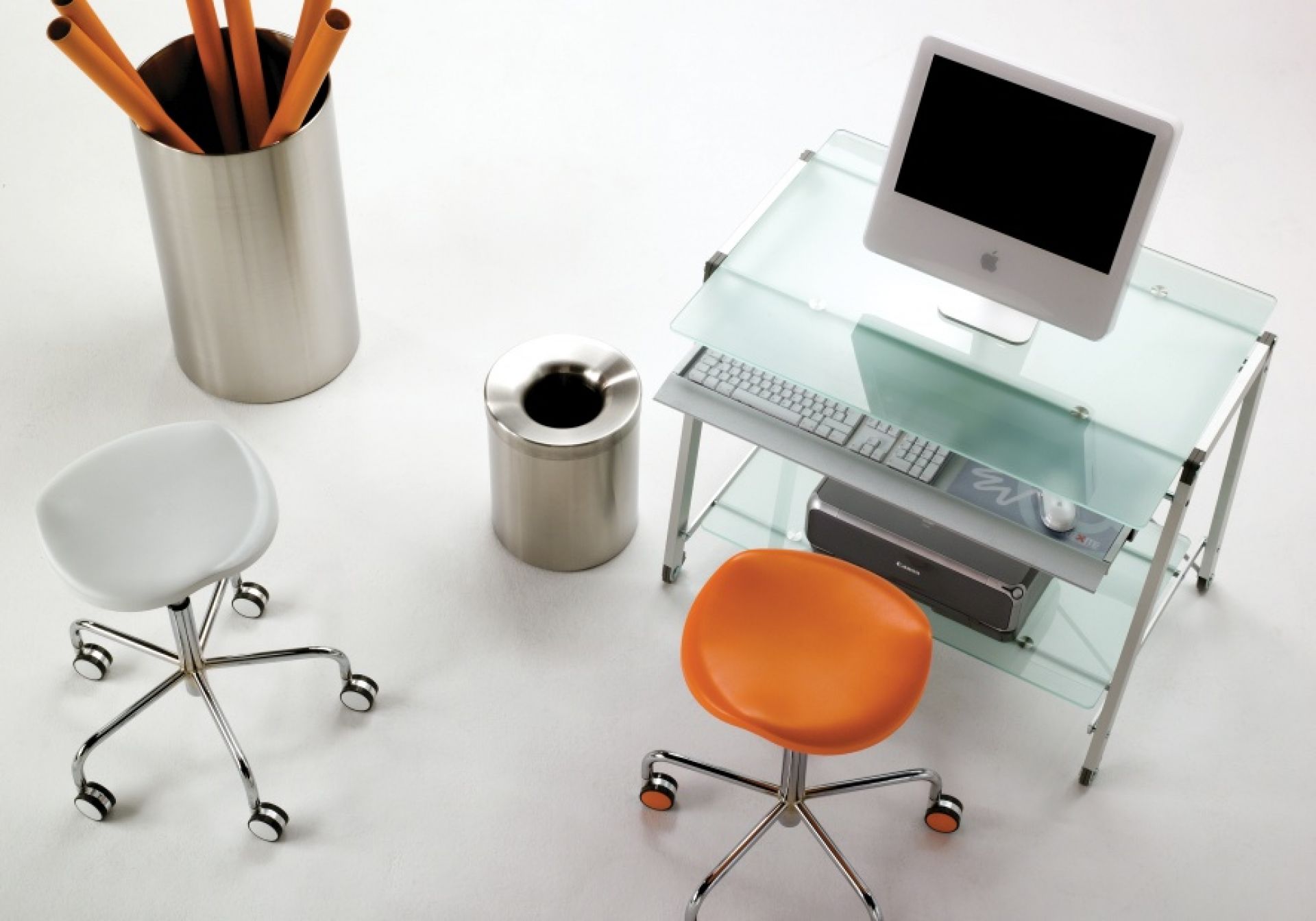 Büro-Setup mit Rexite Welcome Drehhocker in Orange und Weiß, Glas-Schreibtisch und Edelstahl-Accessoires.