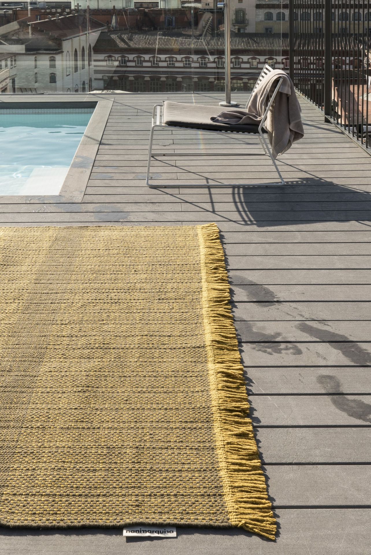 Nanimarquina Tres Outdoor Texture Teppich Senf auf einer Holzterrasse neben einem Pool und Liegestuhl.