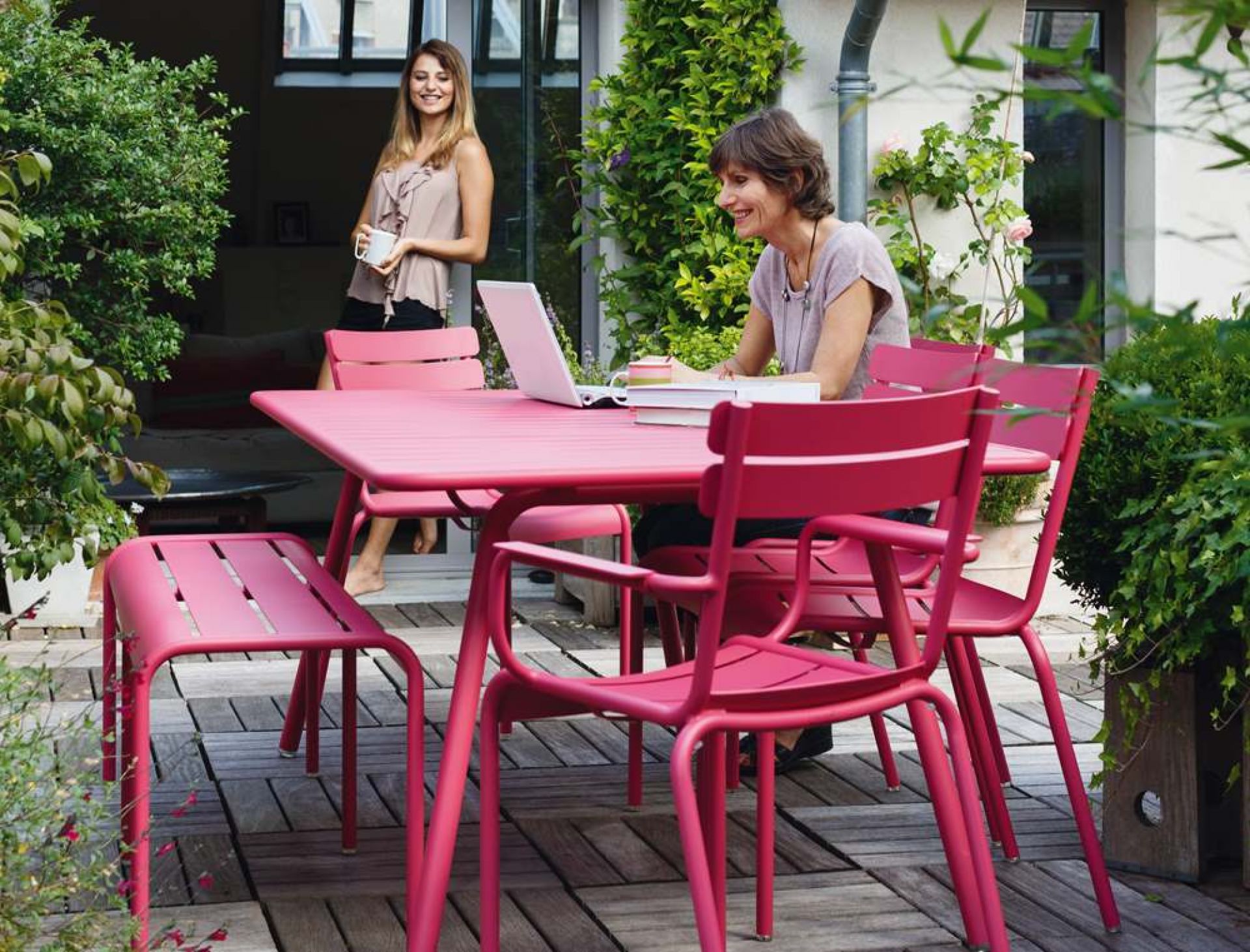 Pinker Fermob Luxembourg Gartentisch mit Vierfußgestell und passenden Stühlen auf einer Holzterrasse.