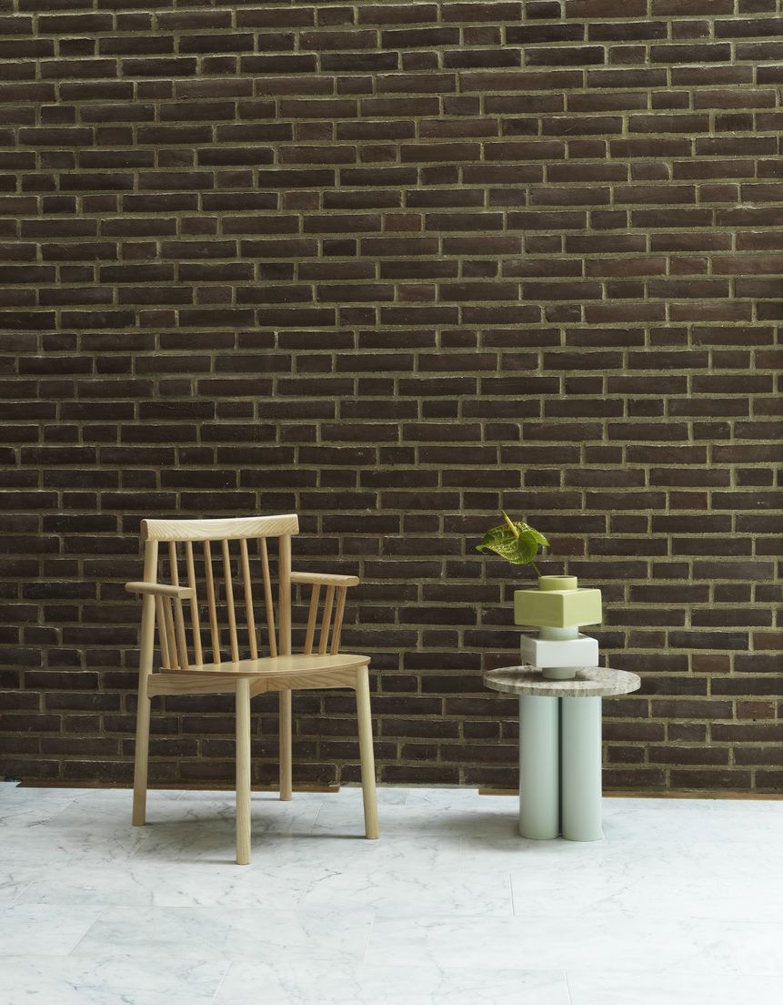 Heller Holzstuhl "Pind Armchair" von Normann Copenhagen mit Armlehnen vor Backsteinwand.