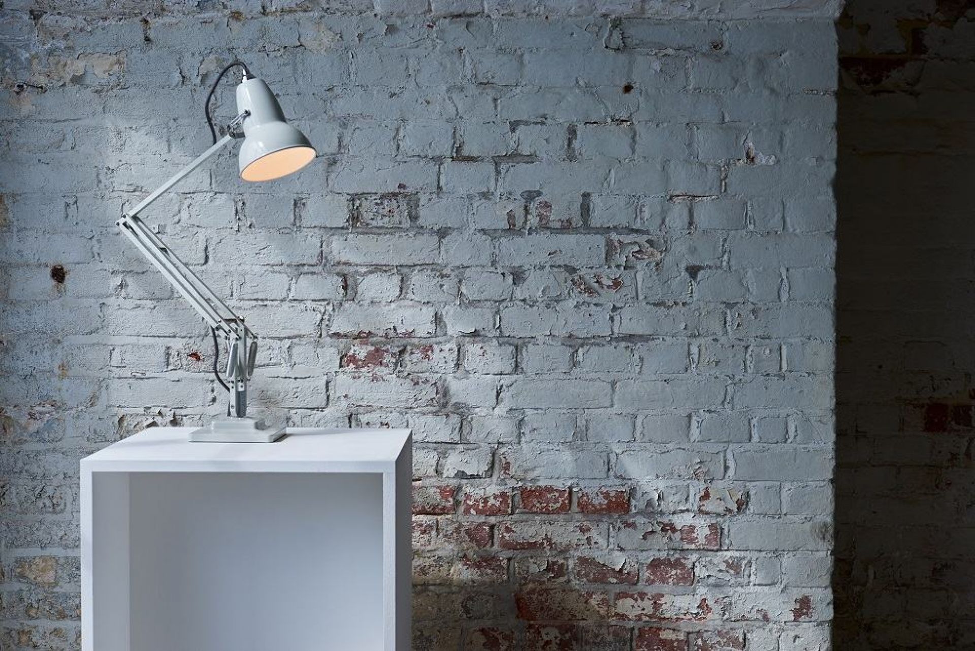 Anglepoise Original 1227 Tischleuchte in Taubengrau auf einem weißen Würfel vor einer Ziegelwand.